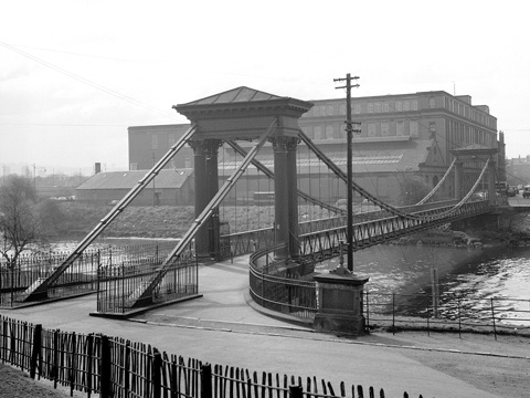 Black and white view of St Andrews Suspension Bridge, a pedestian bridge over the River Clyde. In the background stands a large, industrial building.