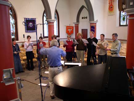 Nigel Swinford conducting nine choir members, grouped in a small floorspace in the middle of the church. Around them are chairs, microphone stands, an overhead projector and a piano