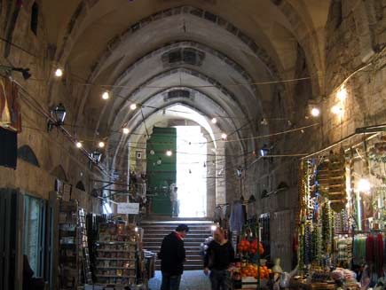 Market stalls are set up in a covered alleyway with a high, arched ceiling. An arched doorway in the background lets some light in, and electric lights are strung across the alley