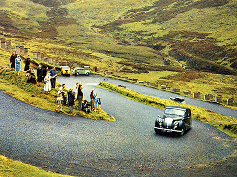Colour image of a vintage Daimler motorcar climbing a steep, double-hairpin section of road in a mountainous area of grass and heather. A small crowd of people stand waving at the roadside.