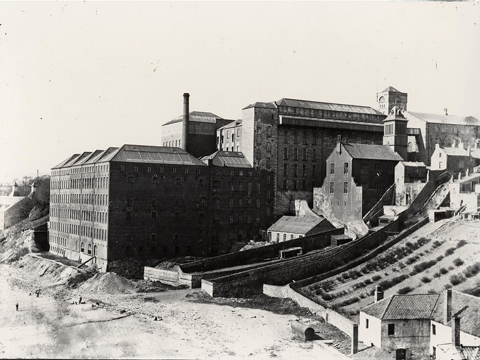 Black and white view of extensive factory building, set on a slope leading down to a beach at the bottom left of frame.
